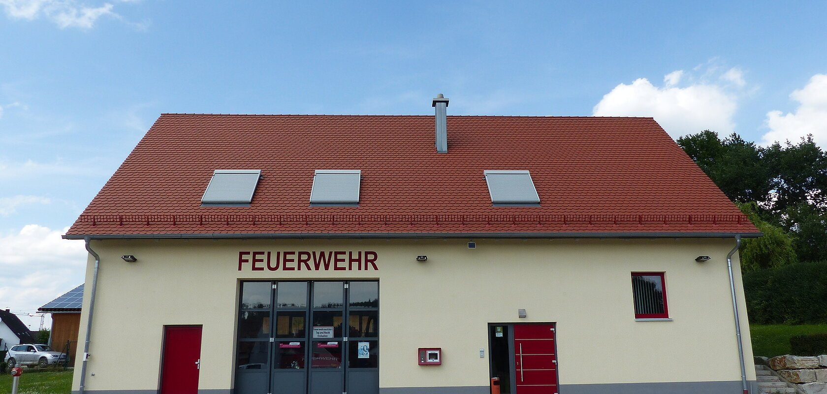 Gelbes Feuerwehrhaus mit rotem Satteldach, drei Dachfenstern und zwei Eingängen und einem Tor. Die Eingangstüren sind beide rot. Um das Gebäude große asphaltierte Fläche. Im Hintergrund blauer Himmel mit Bäumen und einem parkendem Auto.