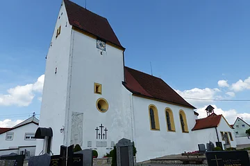 Weiße Pfarrkirche mit Turm, gelb eingerahmten Fenstern, einer Uhr und einem anliegenden Friedhof mit Grabsteinen, unter blauem Himmel.