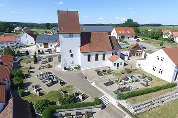 Luftaufnahme einer Kirche mit Friedhof und umliegenden Häusern bei klarem Himmel.