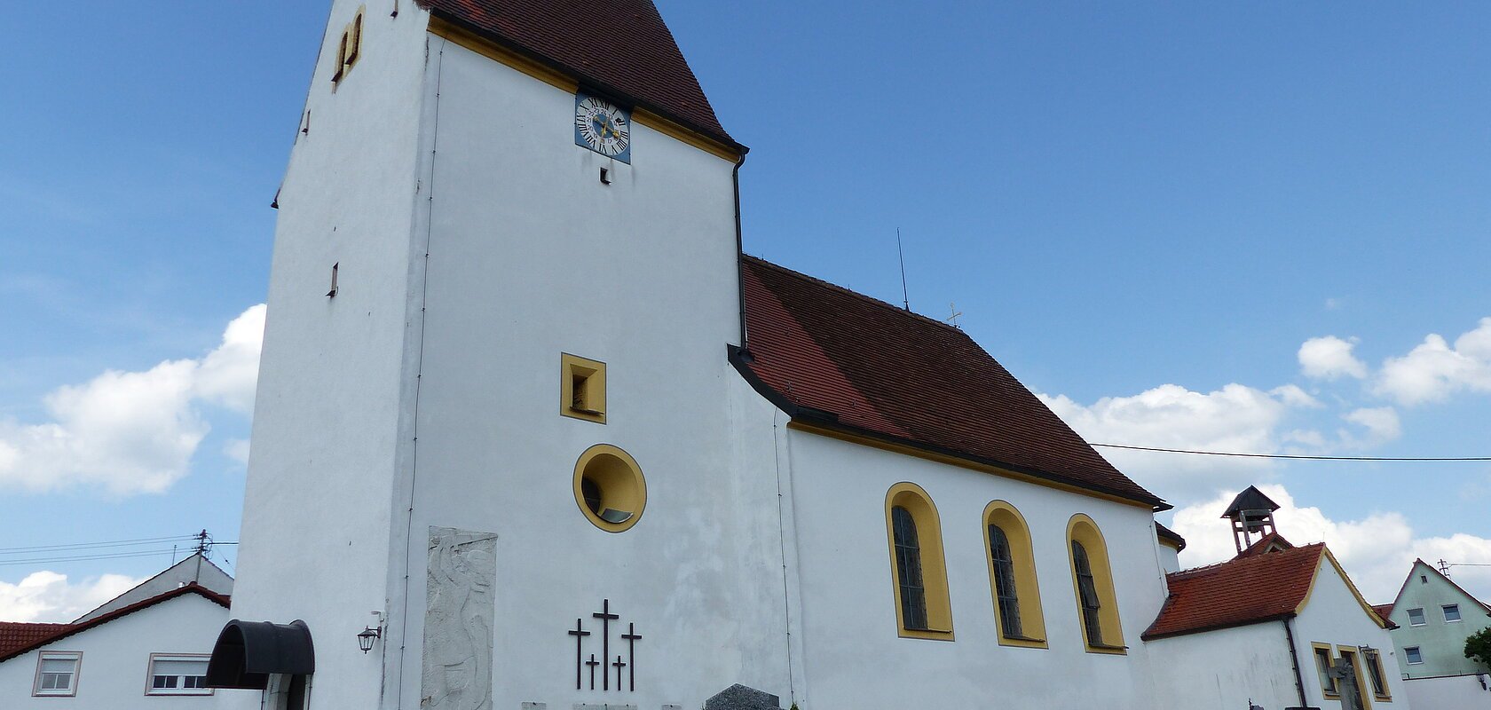 Weiße Pfarrkirche mit Turm, gelb eingerahmten Fenstern, einer Uhr und einem anliegenden Friedhof mit Grabsteinen, unter blauem Himmel.