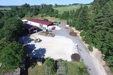 Luftaufnahme eines Forstunternehmens mit Stapeln von Baumstämmen, Gebäuden mit roten Dächern, einer gelben Maschine und einer großen Schotterfläche. Das Forstunternehmen ist umgeben von Bäumen und Feldern.