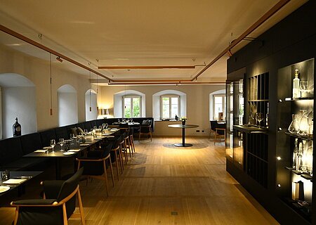 Innenansicht des Restaurants im Hotel by Ferber, in Monheim. Auf der rechten Seite ein großes schwarzes Regal mit verschiedenen Glasbehältern, Weingläsern und Flaschen. Auf der linken Seite schwarze Holztische mit schwarzen Sitzbänken und schwarzen Stühlen zu sehen.