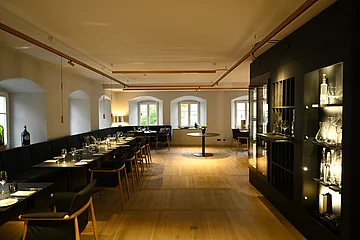 Innenansicht des Restaurants im Hotel by Ferber, in Monheim. Auf der rechten Seite ein großes schwarzes Regal mit verschiedenen Glasbehältern, Weingläsern und Flaschen. Auf der linken Seite schwarze Holztische mit schwarzen Sitzbänken und schwarzen Stühlen zu sehen.