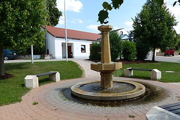 Im Vordergrund ein steinerner Brunnen mit Wasser, auf gepflastertem Platz, umgeben von mehreren Bänken, Bäumen und einem weißen Gebäude mit rotem Dach im Hintergrund. Strahlend blauer Himmel mit vereinzelten Wolken.