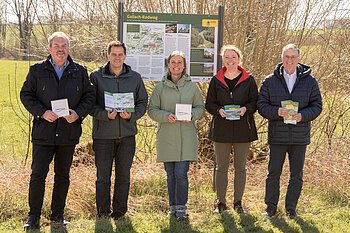 Fünf Personen stehen lächelnd vor einer Informationstafel zum Gailach-Radweg in der Monheimer Alb und halten Broschüren und Schilder in der Hand, umgeben von grüner Wiese und Bäumen.