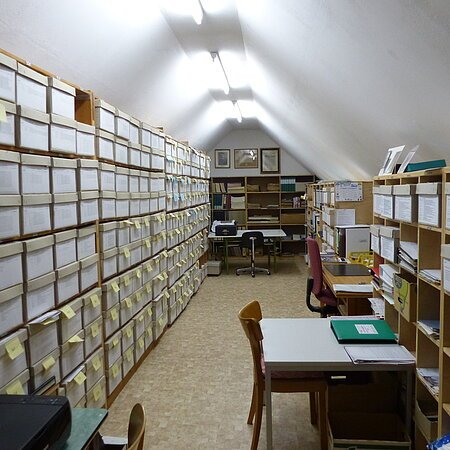 Archivraum mit Regalen voller beschrifteter Kartons und vier Tischen mit vier Stühlen unter einer schrägen weißen Decke mit Lichtröhren.