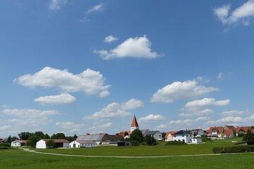 Dorf mit Kirche, Häusern und einer Feldstraße vor grünem Feld unter blauem Himmel mit weißen Wolken.