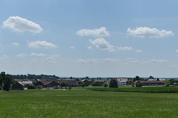 Blick auf eine grüne Wiese im Vordergrund, mit Wohnhäusern und Bäumen unter blauem Himmel mit vereinzelten Wolken im Hintergrund.