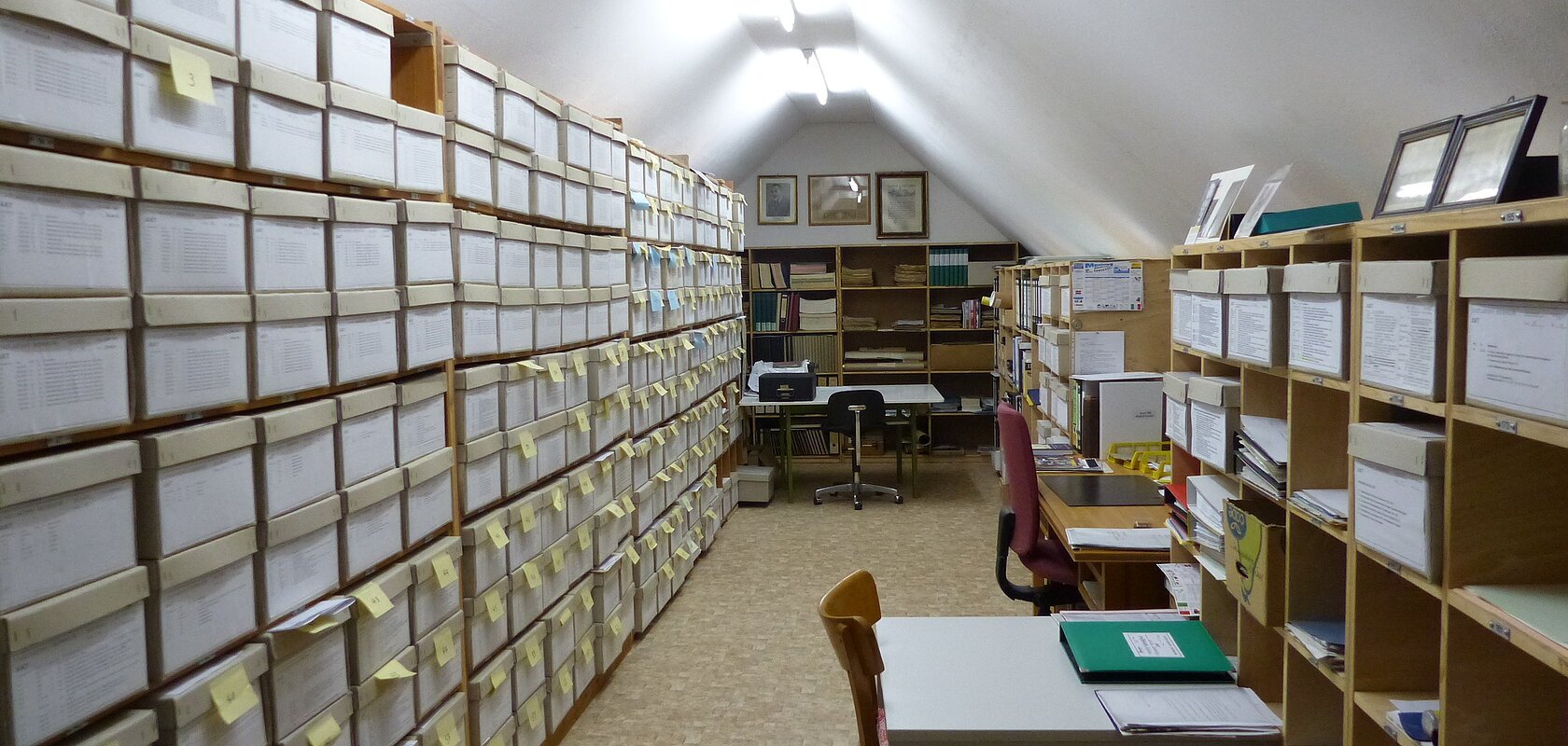 Archivraum mit Regalen voller beschrifteter Kartons und vier Tischen mit vier Stühlen unter einer schrägen weißen Decke mit Lichtröhren.