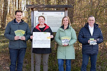 Vier Personen stehen lächelnd vor einer Informationstafel im Wald und halten Broschüren und eine Karte in den Händen.