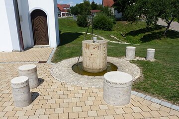 Brunnen mit zylindrischem Steinbecken mit laufendem Wasser und einem gepflastertem Rundplatz außenrum. Fünf weitere beschriftete zylindrische Steinsäulen auf gepflastertem Platz vor weißem Gebäude und grüner Wiese.