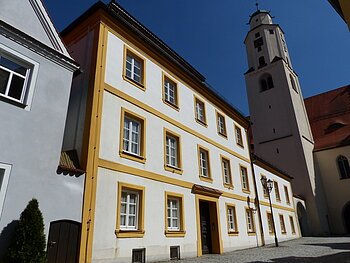 Dreistöckiges weißes Gebäude mit gelben Fensterrahmen und angrenzendem Kirchturm auf einem gepflastertem Platz, unter blauem Himmel.