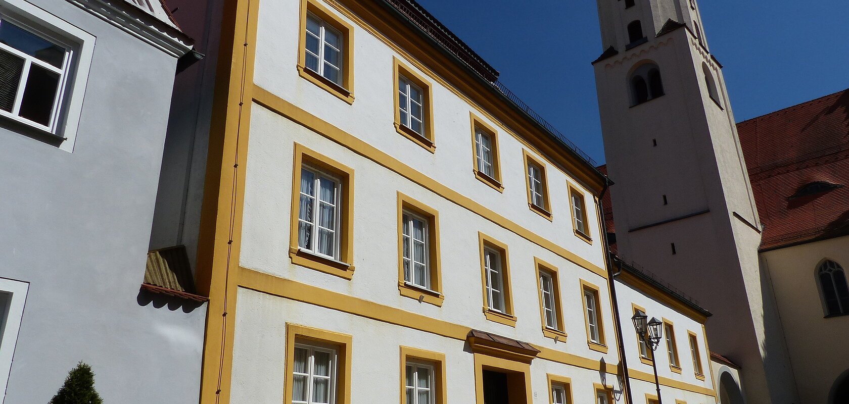 Dreistöckiges weißes Gebäude mit gelben Fensterrahmen und angrenzendem Kirchturm auf einem gepflastertem Platz, unter blauem Himmel.