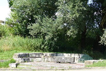 Rastplatz für Radler in Form von einer steinernen Sitzbank aus Stufen vor dichten grünen Bäumen und Sträuchern im Freien bei Tageslicht. Im Vordergrund ein Stück Straße zu sehen.