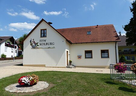 Ein gelbes Gebäude, das Feuerwehrhaus in Kölburg mit rotem Dach, vier Fenstern und einem Defibrillator. Auf dem Gebäude die Aufschrift "FFW Kolburg", davor ein Blumenbeet auf einer Rasenfläche und ein weiterer Blumenkasten auf einem Geländer. Im Hintergrund eine Straße mit einem weißen Haus.