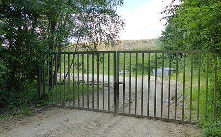Verschlossenes Metalltor auf einem unbefestigten Weg, umgeben von Bäumen und Gras. Im Hintergrund ist ein Müllcontainer und ein Hügel sichtbar.