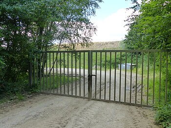Verschlossenes Metalltor auf einem unbefestigten Weg, umgeben von Bäumen und Gras. Im Hintergrund ist ein Müllcontainer und ein Hügel sichtbar.
