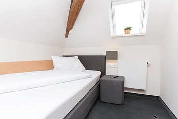Einzelzimmer in einem Hotel mit einem Einzelbett links an der weißen Wand, einem grauem Hocker rechts daneben, auf dem eine  Fernbedienung liegt und einer grauen Leuchte oberhalb an der Wand. Dachschräge mit Holzbalken, rechts davon und kleines Fenster mit Pflanze auf dem Fensterbrett. Unter dem Fester eine kleine weiße Heizung an der Wand angebracht.