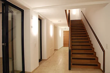 Flur mit beleuchteten weißen Wänden durch Wandleuchten links im Bild und einer Glastür. Eine Holztreppe, die nach oben führt rechts im Bild. Hinten im Gang eine helle Holztür, heller Bodenbelag aus Fließen.