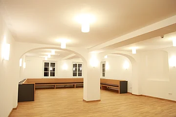 Großer leerstehender heller Raum mit hellem Holzfußboden, weißen Wänden, Holzbänken hinten entlang der Wände, mehreren Deckenlampen und Rundbögen in der Mitte des Raums.