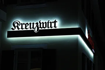 Zwei weiß beleuchtete dunkelrote Schriftzüge „Kreuzwirt“ an einer helle Hauswand bei Nacht um die Ecke verbunden mit einem dunkelrotem Balken darunter.