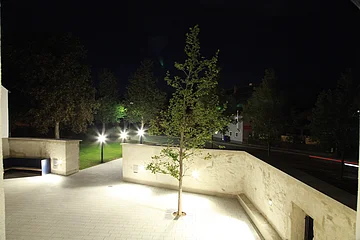 Beleuchteter Innenhof bei Nacht, eingezäunt mit einer hohe weißen Mauer, darin ein Baum rechts im Bild und eine an der Wand befestigte Sitzbank links im Bild. Außerhalb drei leuchtende Laternen am Gehweg entlang, Bäume auf grüner Wiese und im Hintergrund eine Straße mit einem grauen Haus.