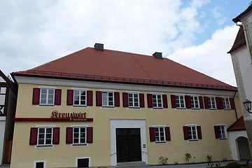 Gelbes zweistöckiges Gebäude mit vielen Fenstern mit jeweils zwei roten Fensterläden an jedem Fenster, einem rotem Ziegeldach und dem Schriftzug „Kreuzwirt - Haus der Kultur“ links vom Eingang. In der Mitte eine große braune zweiflüglige Holztür. Vor dem Gebäude eine Straße, im Hintergrund blauer Himmel mit Wolken.