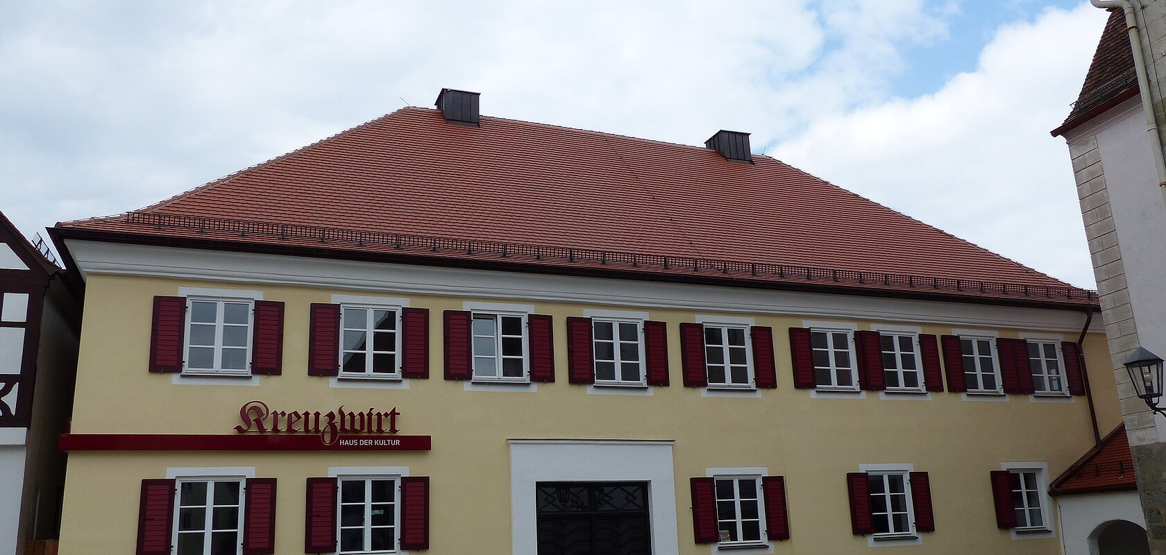 Gelbes zweistöckiges Gebäude mit vielen Fenstern mit jeweils zwei roten Fensterläden an jedem Fenster, einem rotem Ziegeldach und dem Schriftzug „Kreuzwirt - Haus der Kultur“ links vom Eingang. In der Mitte eine große braune zweiflüglige Holztür. Vor dem Gebäude eine Straße, im Hintergrund blauer Himmel mit Wolken.