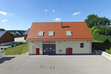 Feuerwehrgebäude mit der Aufschrift "Feuerwehr" in rot, mit rotem Dach, drei Dachfenstern und zwei roten Türen, sowie einem großen grauen Tor. Davor großer gepflasterter Hof im Vordergrund. Im Hintergrund Wohnhäuser, grüne Bäume und blauer Himmel.