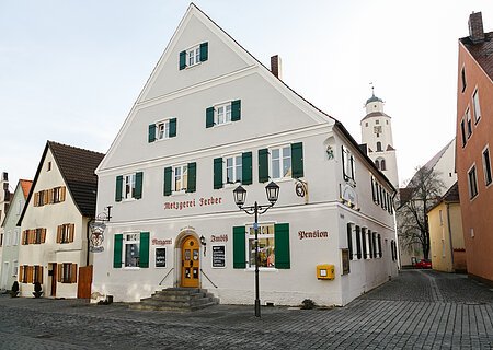 Graues mehrstöckiges Gebäude mit grünen Fensterläden, einer Treppe mit Geländern und dem Schriftzug "Metzgerei Ferber" oberhalb der Eingangstür, an einer gepflasterten Straße. Links von der Tür der Schriftzug "Metzgerei", rechts davon "Imbiss" und "Pension" an der Fassade. Ein gelber Briefkasten hängt an der vorderen Fassade.