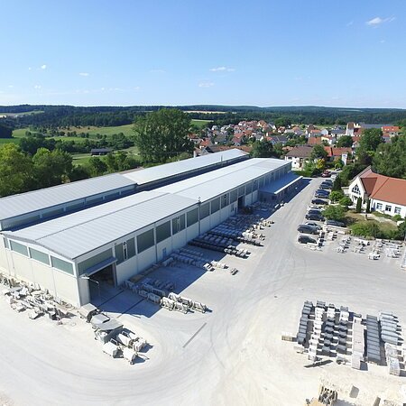 Luftaufnahme eines Industriegebäudes von einem Natursteinwerk mit asphaltierten Lagerflächen und Fahrzeugen darauf. Ein Dorf, grüne Landschaft und blauer Himmel im Hintergrund.