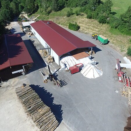 Luftaufnahme eines Holzlagers mit zwei großen Gebäuden, Holzstapeln und Maschinen auf Asphaltfläche, umgeben von Wiese und grünen Bäumen.