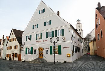 Graues mehrstöckiges Gebäude mit grünen Fensterläden, einer Treppe mit Geländern und dem Schriftzug "Metzgerei Ferber" oberhalb der Eingangstür, an einer gepflasterten Straße. Links von der Tür der Schriftzug "Metzgerei", rechts davon "Imbiss" und "Pension" an der Fassade. Ein gelber Briefkasten hängt an der vorderen Fassade.