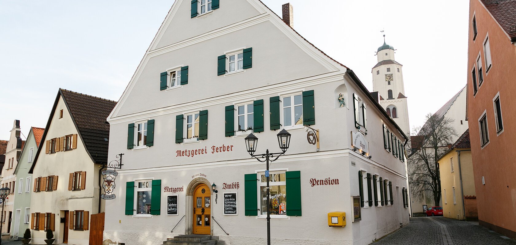 Graues mehrstöckiges Gebäude mit grünen Fensterläden, einer Treppe mit Geländern und dem Schriftzug "Metzgerei Ferber" oberhalb der Eingangstür, an einer gepflasterten Straße. Links von der Tür der Schriftzug "Metzgerei", rechts davon "Imbiss" und "Pension" an der Fassade. Ein gelber Briefkasten hängt an der vorderen Fassade.