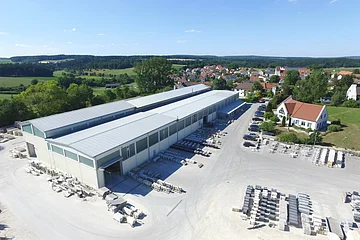 Luftaufnahme eines Industriegebäudes von einem Natursteinwerk mit asphaltierten Lagerflächen und Fahrzeugen darauf. Ein Dorf, grüne Landschaft und blauer Himmel im Hintergrund.