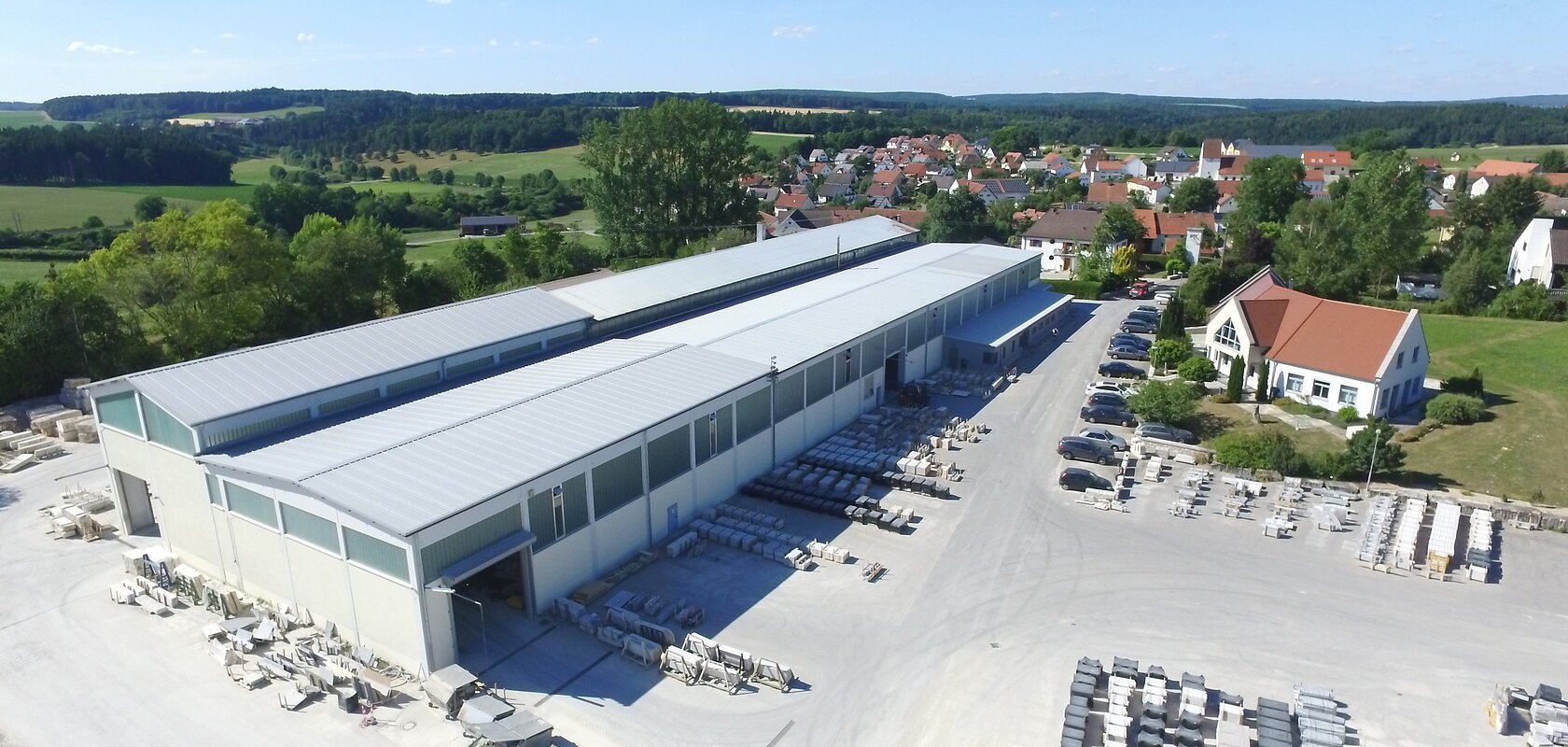 Luftaufnahme eines Industriegebäudes von einem Natursteinwerk mit asphaltierten Lagerflächen und Fahrzeugen darauf. Ein Dorf, grüne Landschaft und blauer Himmel im Hintergrund.