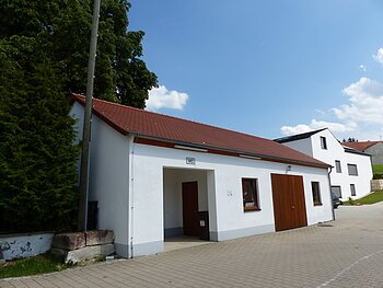 Weißes längliches Gebäude mit rotem Dach und WC-Schild über dem Eingang. Rechts vom Eingang ein Fenster, ein Holztor und weiteres Fenster daneben. Links vom Gebäude sind Bäume, vorderhalb ein gepflasterter Platz,  rechts im Bild ein Wohnhaus. Im Hintergrund strahlend blauer Himmel.