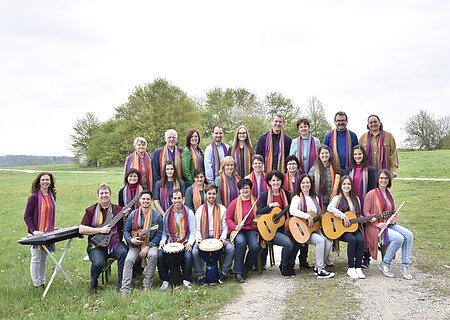 De Lumina - der Chor Gruppenfoto von 28 Personen mit Musikinstrumenten und bunten Schals um den Hals auf einer Wiese vor Bäumen bei bewölktem Himmel.
