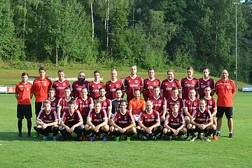 Fußballmannschaft mit 29 Spielern und Trainern in roten Trikots auf grünem Spielfeld vor Bäumen posierend.