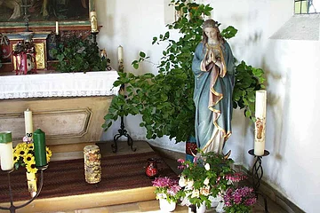 Innenansicht einer kleinen Kapelle mit der Statue einer betenden Frau mit blauem Mantel in einer Ecke umgeben von Kerzen, Pflanzen und Blumen in einem weißen Raum. Links im Bild ein geschmückter Altar.