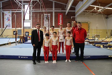 Gruppenfoto mit sieben Jungen in Turnkleidung, zwei Männern in Sportkleidung rechts von ihnen und einem Mann im Anzug links, in einer Turnhalle auf blauer Matte vor Turngeräten.