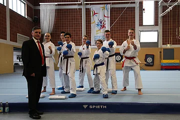Gruppe von sieben Karatekämpfern in weißen Anzügen, teilweise mit blauen Handschuhen auf blauer Matte und einem Mann im Anzug links von der Gruppe, in einer Sporthalle. Im Hintergrund ein Banner und weitere Sportgeräte.