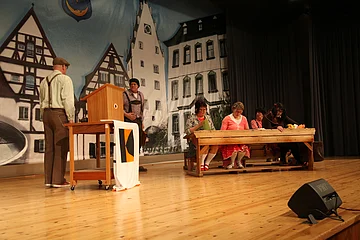 Theaterszene mit sechs Personen auf der Bühne: zwei Männer stehen neben einem Rednerpult und schauen sich an, drei Frauen sitzen am Holztisch daneben und eine weitere Person sitz im Hintergrund. Kulisse mit Fachwerkhäusern und ein schwarzer Lichtstrahler vorne auf der Bühne.