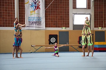 Sieben Turnerinnen in farbigen Anzügen führen eine Partnerakrobatik-Übung auf einer blauen Matte in einer Sporthalle durch. Im Hintergrund weitere Sportgeräte und ein Banner mit dem Titel "Internationales Deutsches Turnfest Berlin 03. - 10-Juni 2017" an der Wand.