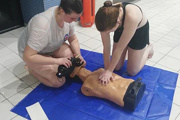 Zwei Frauen üben Herz-Lungen-Wiederbelebung an einer Übungspuppe auf einer blauen Matte in einem Schwimmbad.