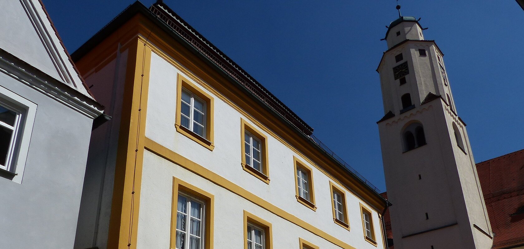 Mehrstöckiges weißes Gebäude mit gelben Fenster- und Türrahmen und gelben durchgehenden Balken über das ganze Gebäude. Angrenzend an eine Kirche der Kirchturm zwischen den beiden Gebäuden bei blauem Himmel.