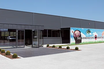 Eingang eines modernen grauen Gebäudes mit Glasfront, Flachdach und Werbebannern an der Fassade, bei strahlend blauem Himmel. Vor dem Eingang zwei bepflanzte Beete links und rechts, umgeben vom großen gepflastertem Hof.