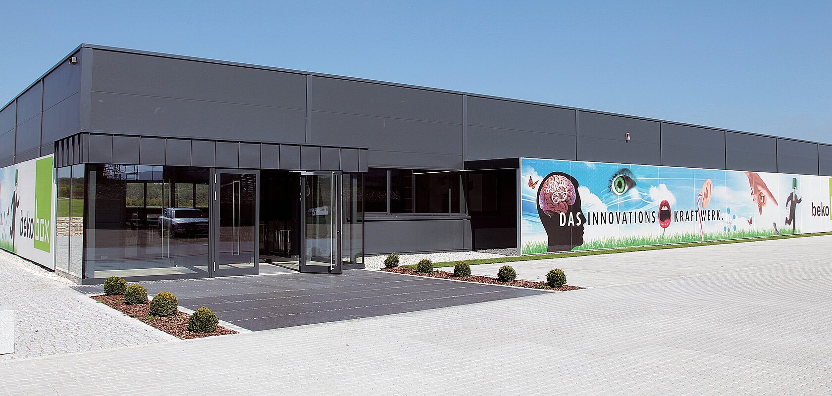Eingang eines modernen grauen Gebäudes mit Glasfront, Flachdach und Werbebannern an der Fassade, bei strahlend blauem Himmel. Vor dem Eingang zwei bepflanzte Beete links und rechts, umgeben vom großen gepflastertem Hof.