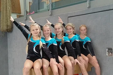 Fünf Mädchen in Turnanzügen sitzen auf einem Turngerät in einer Sporthalle und strecken ihre Arme nach oben und unten aus.