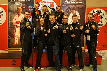 Neun Personen in Sportkleidung posieren mit geballten Fäusten vor Werbebannern zum Karatesport und einem Sieger-Podest.